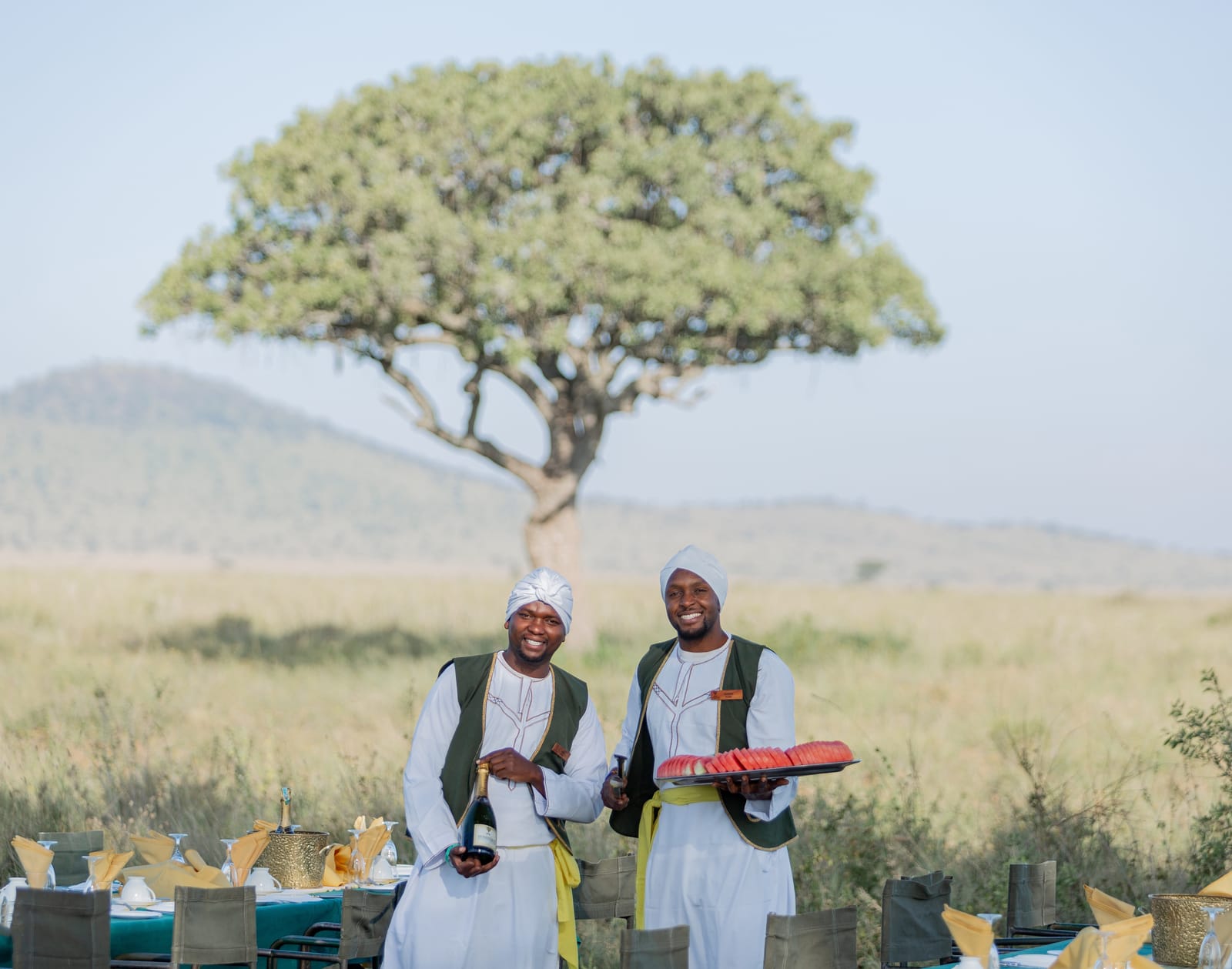 Two hosts welcoming guests to a bush breakfast under an acacia